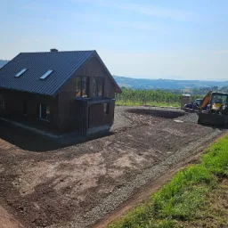 Nowoczesny dom z ciemnego drewna z metalowym dachem i oknami dachowymi, teren wokół domu przygotowany pod trawnik, widoczny wykop i utwardzona droga dojazdowa, w tle krajobraz z drzewami i wzgórzami.