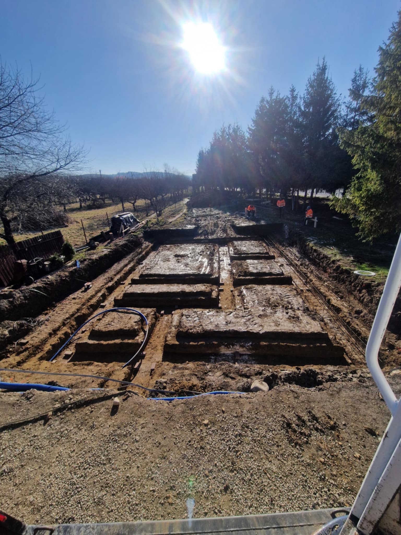 Przygotowany wykop pod fundamenty z częściowo ułożonym zbrojeniem, widoczne niebieskie rury instalacyjne, w tle drzewa i błękitne niebo z silnym słońcem.