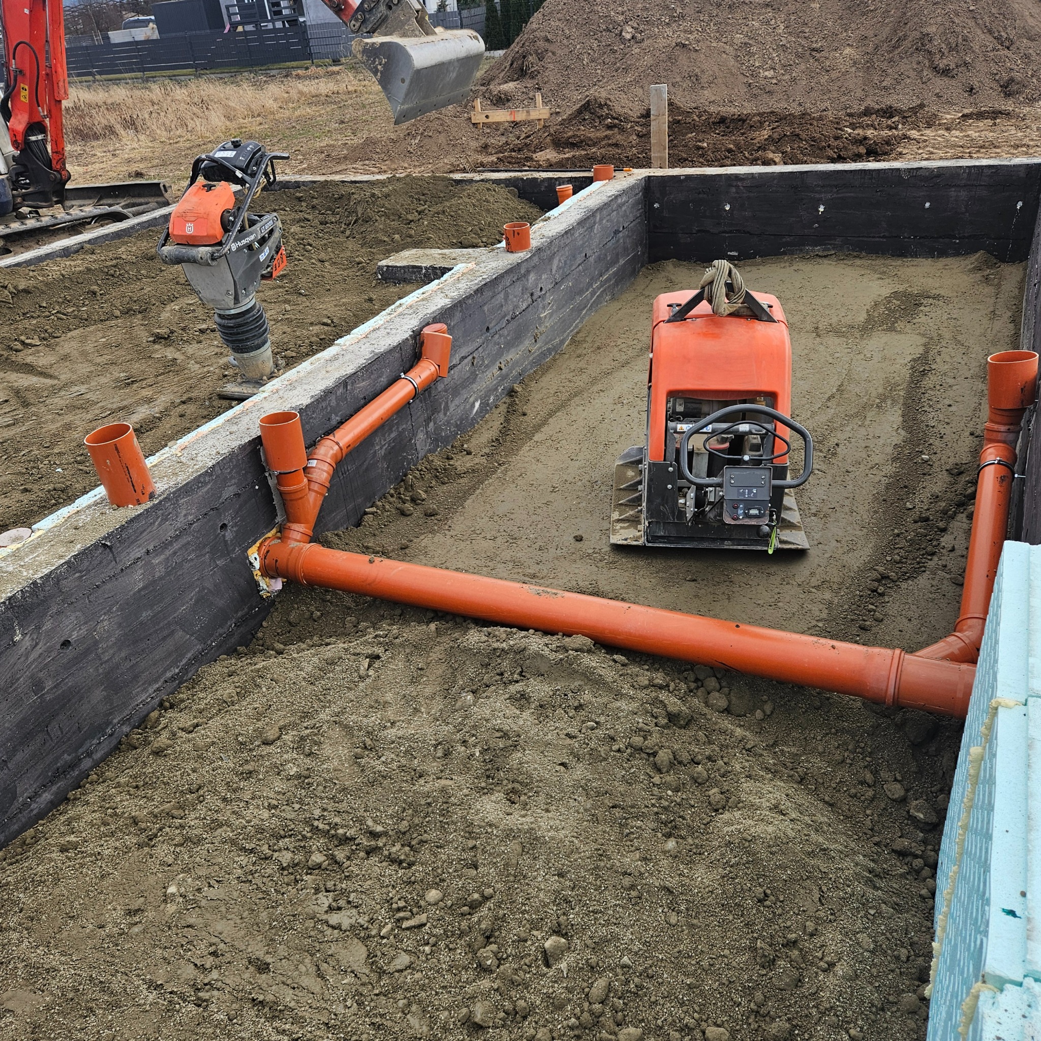 Wykop pod fundamenty z ułożonymi rurami drenażowymi i zagęszczarkami wibracyjnymi, widoczna izolacja pionowa fundamentów i przygotowany teren pod wylewkę.