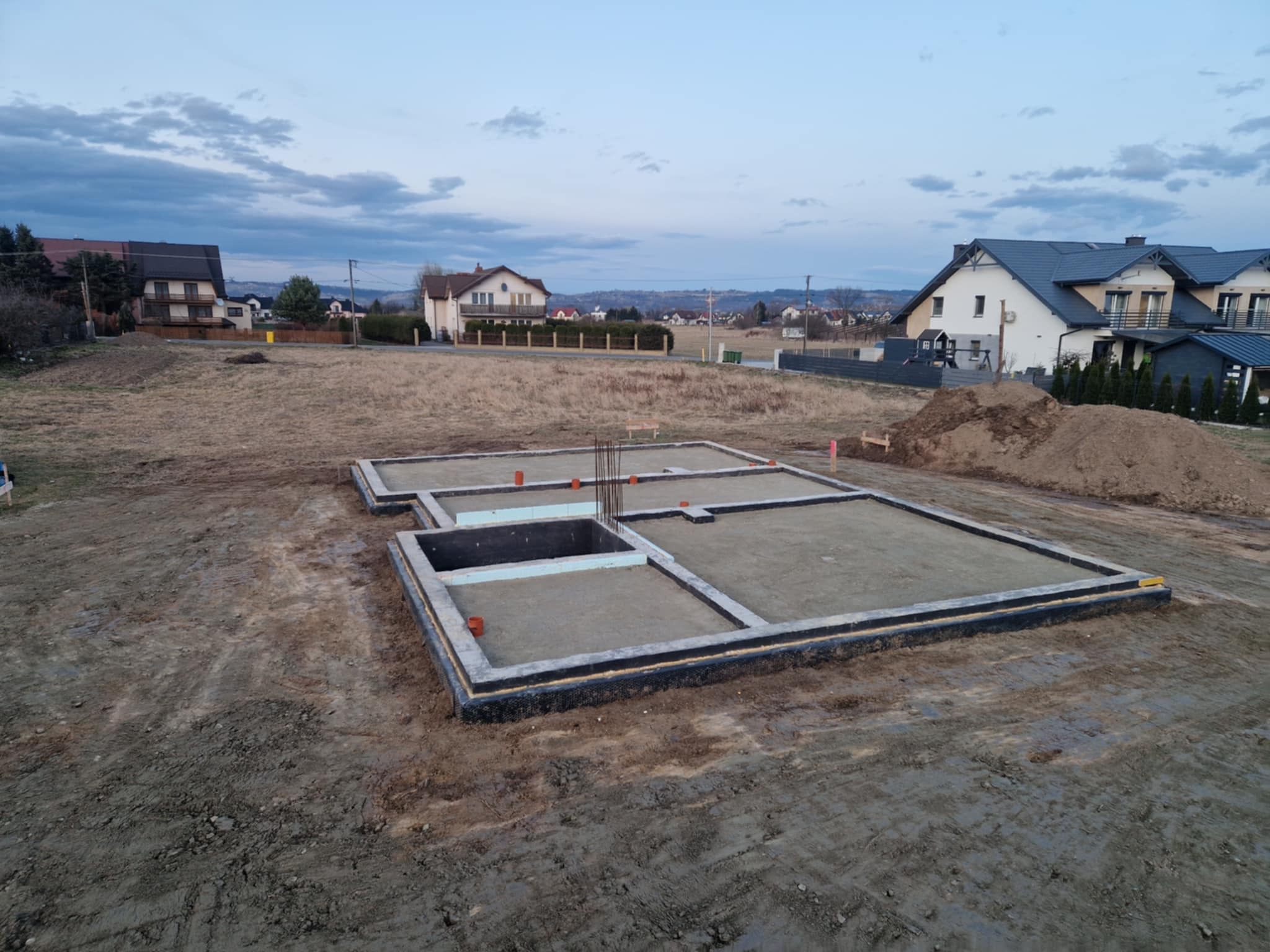 Zalane fundamenty domu jednorodzinnego z widoczną izolacją przeciwwilgociową i wystającymi prętami zbrojeniowymi na tle krajobrazu z zabudową mieszkalną.
