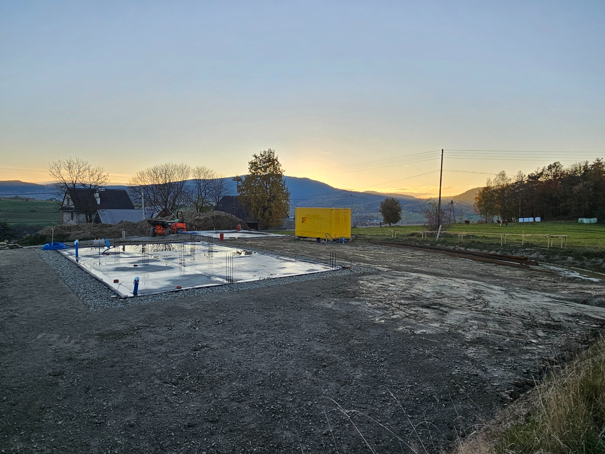 Wylany fundament z wystającymi prętami zbrojeniowymi na przygotowanym terenie, widoczny żółty kontener budowlany i krajobraz z górami w tle o zachodzie słońca.
