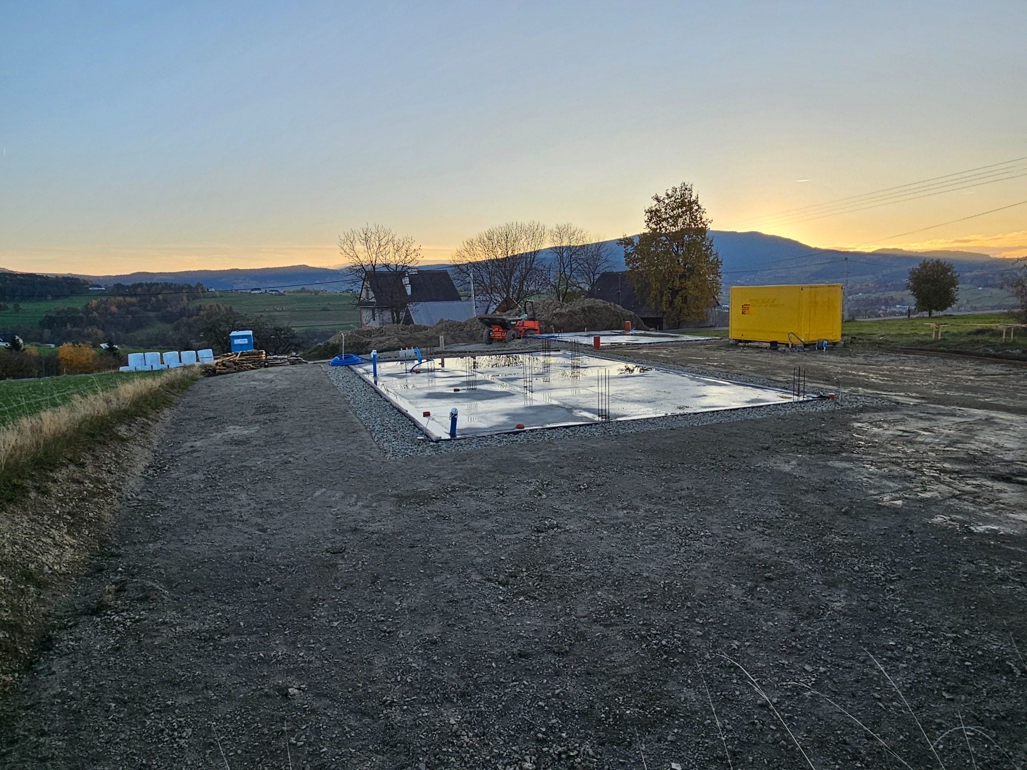 Przygotowany teren pod fundamenty z widocznym zbrojeniem, zalany betonem, otoczony żwirem, z widokiem na pagórkowaty krajobraz o zachodzie słońca.