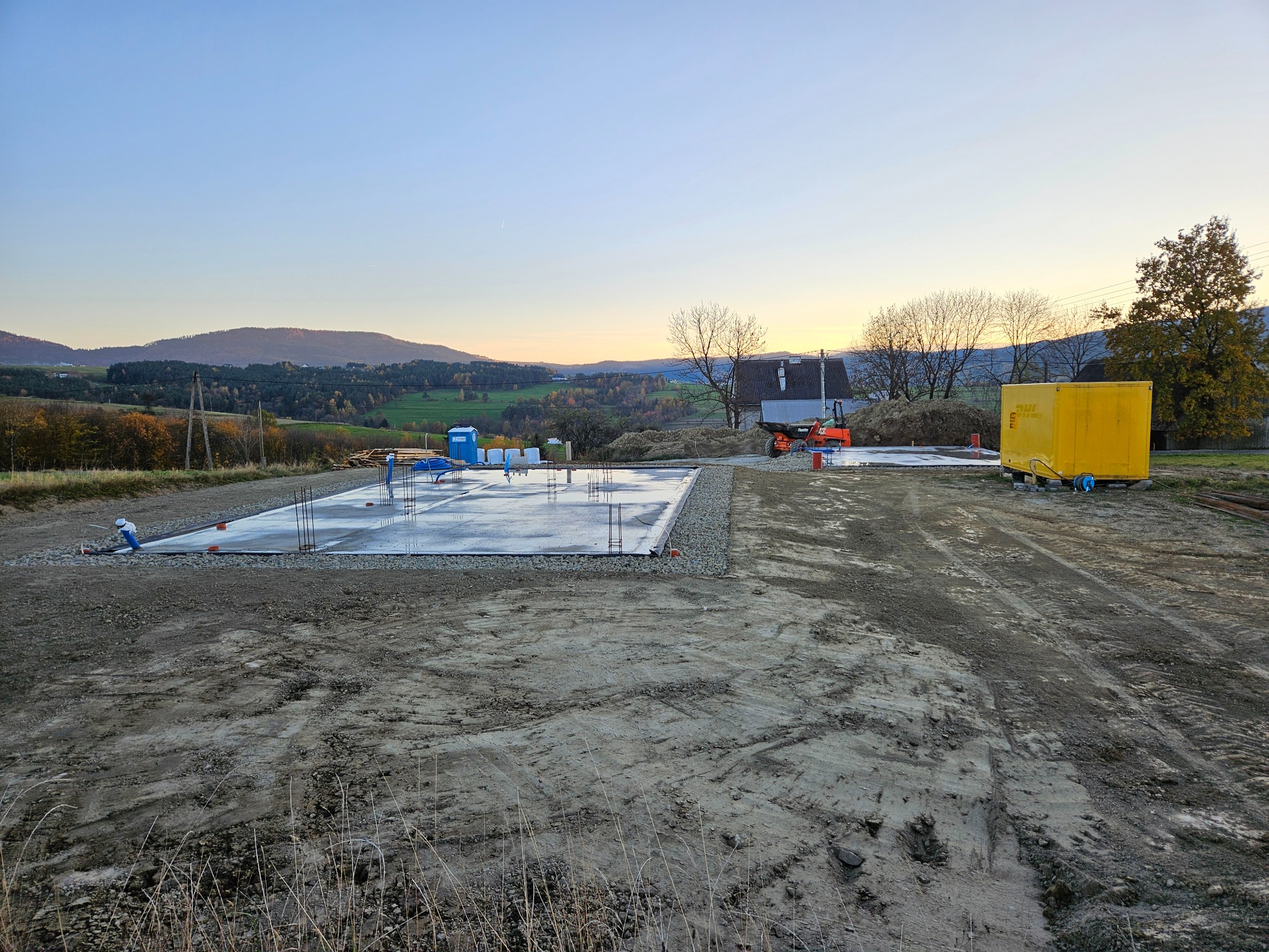 Wylana płyta fundamentowa na przygotowanym terenie, widoczne zbrojenia, w tle krajobraz pagórkowaty o zmierzchu, na placu budowy żółty kontener i sprzęt budowlany.