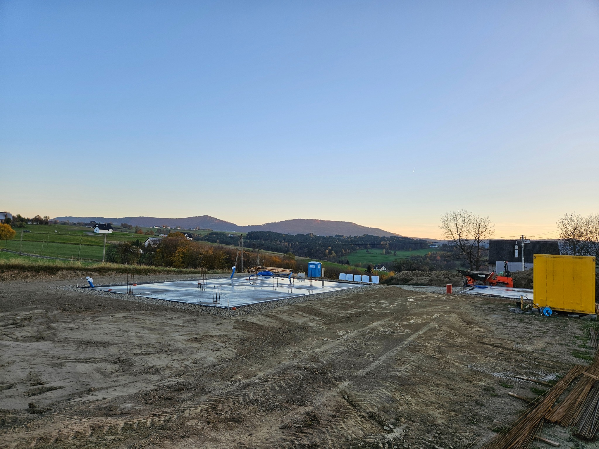Wylana płyta fundamentowa z wystającymi prętami zbrojeniowymi, widoczna ubikacja TOI TOI, mini wywrotka i teren przygotowany pod budowę na tle pagórkowatego krajobrazu o zmierzchu.