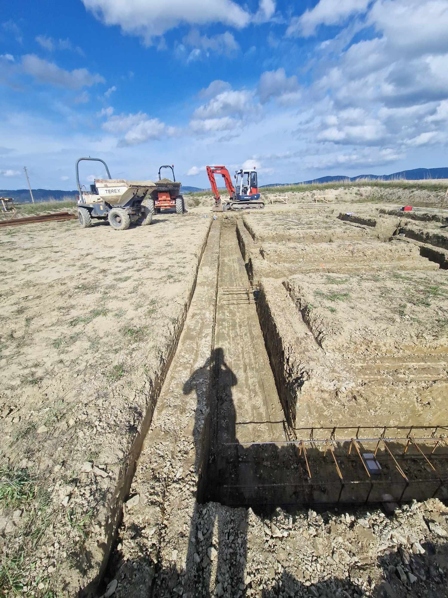 Przygotowanie terenu pod fundamenty: wykopane rowy z widocznym zbrojeniem, na drugim planie maszyny budowlane - wywrotka i mini koparka, błękitne niebo z chmurami w tle.