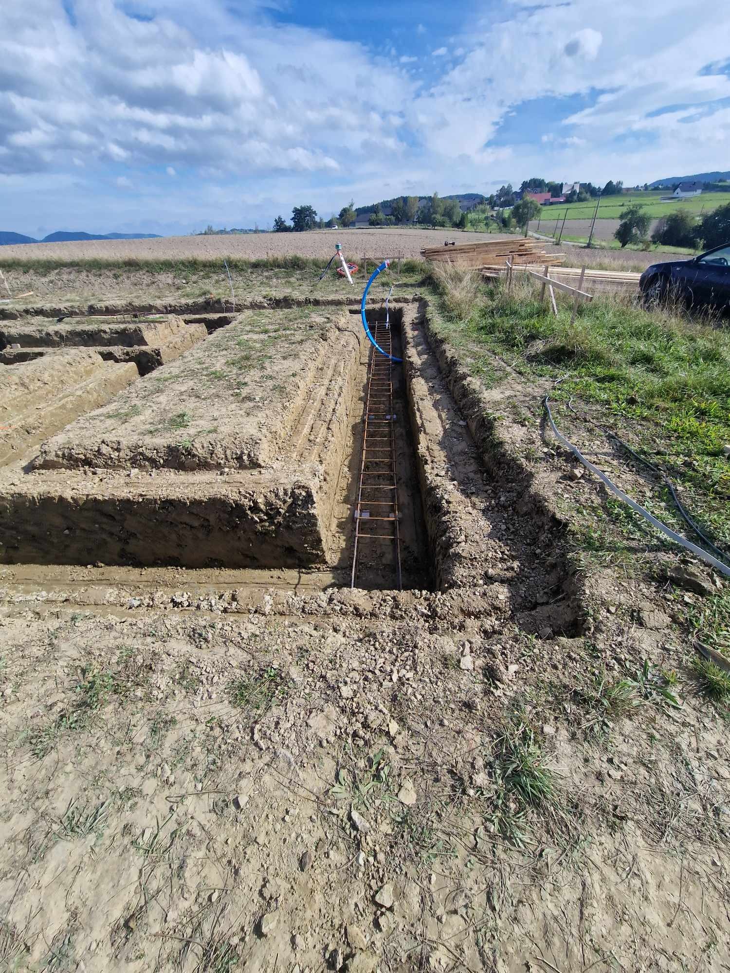 Szeroki kadr na wykopane rowy pod fundamenty z widocznym zbrojeniem, niebieską rurą i stertą drewna w tle na tle krajobrazu.
