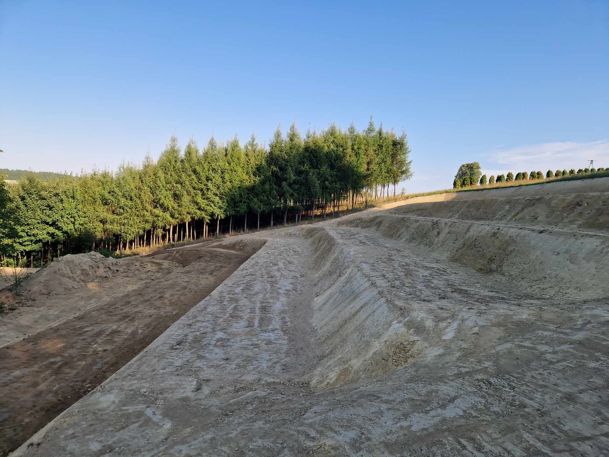 Wyprofilowany teren z rowem, świeżo po pracach ziemnych, z widocznymi śladami opon i rzędem drzew w tle pod błękitnym niebem.
