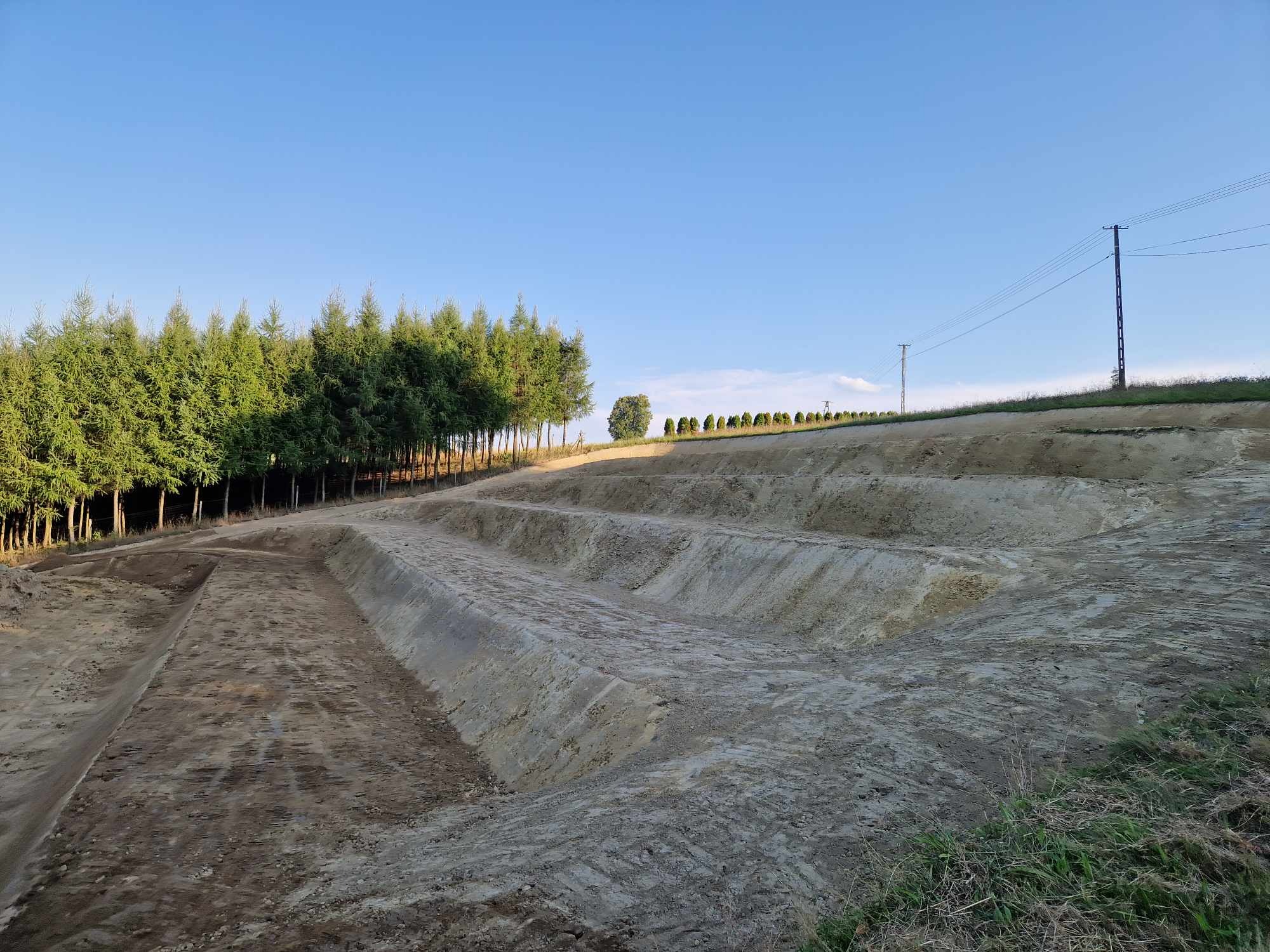 Wielopoziomowe wykopy w ziemi, widoczne ślady opon, w tle rząd drzew iglastych i słupy energetyczne na tle błękitnego nieba.