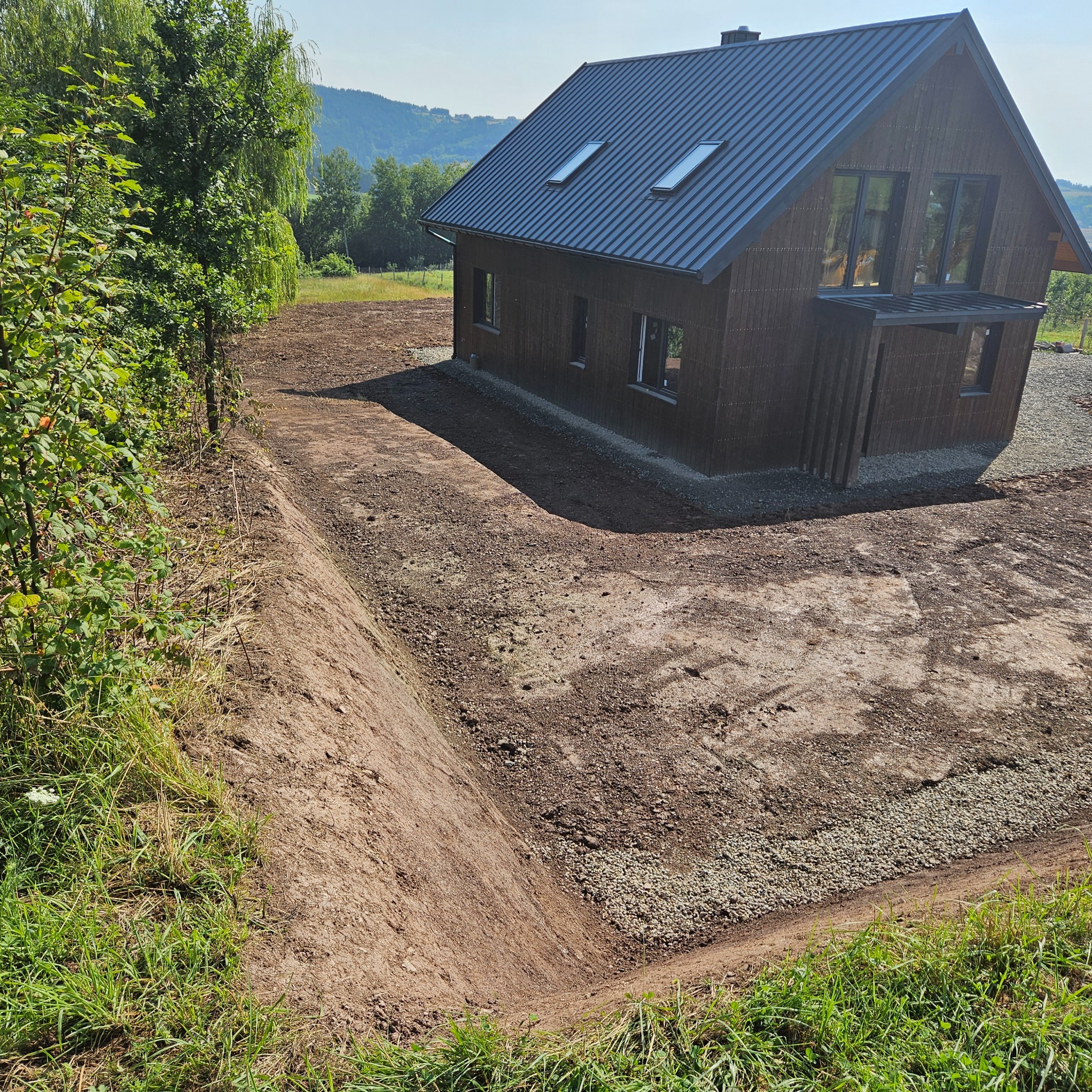Przygotowany teren pod budowę domu z widocznym wykopem, częściowo utwardzoną drogą dojazdową i nowoczesnym budynkiem w tle.