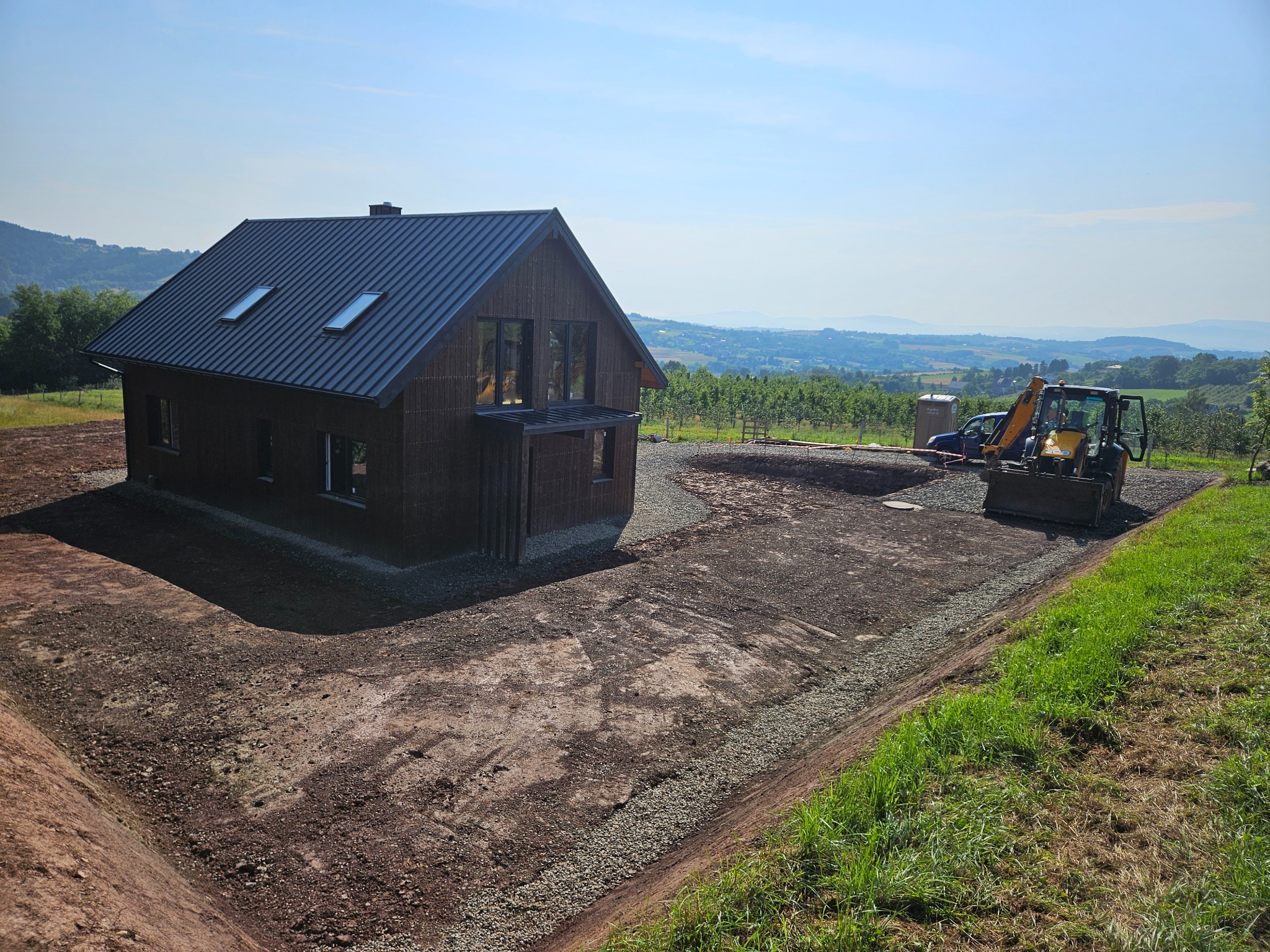 Nowoczesny dom z ciemnego drewna z metalowym dachem i oknami dachowymi, teren wokół domu przygotowany pod trawnik, widoczny wykop i utwardzona droga dojazdowa, w tle krajobraz z drzewami i wzgórzami.