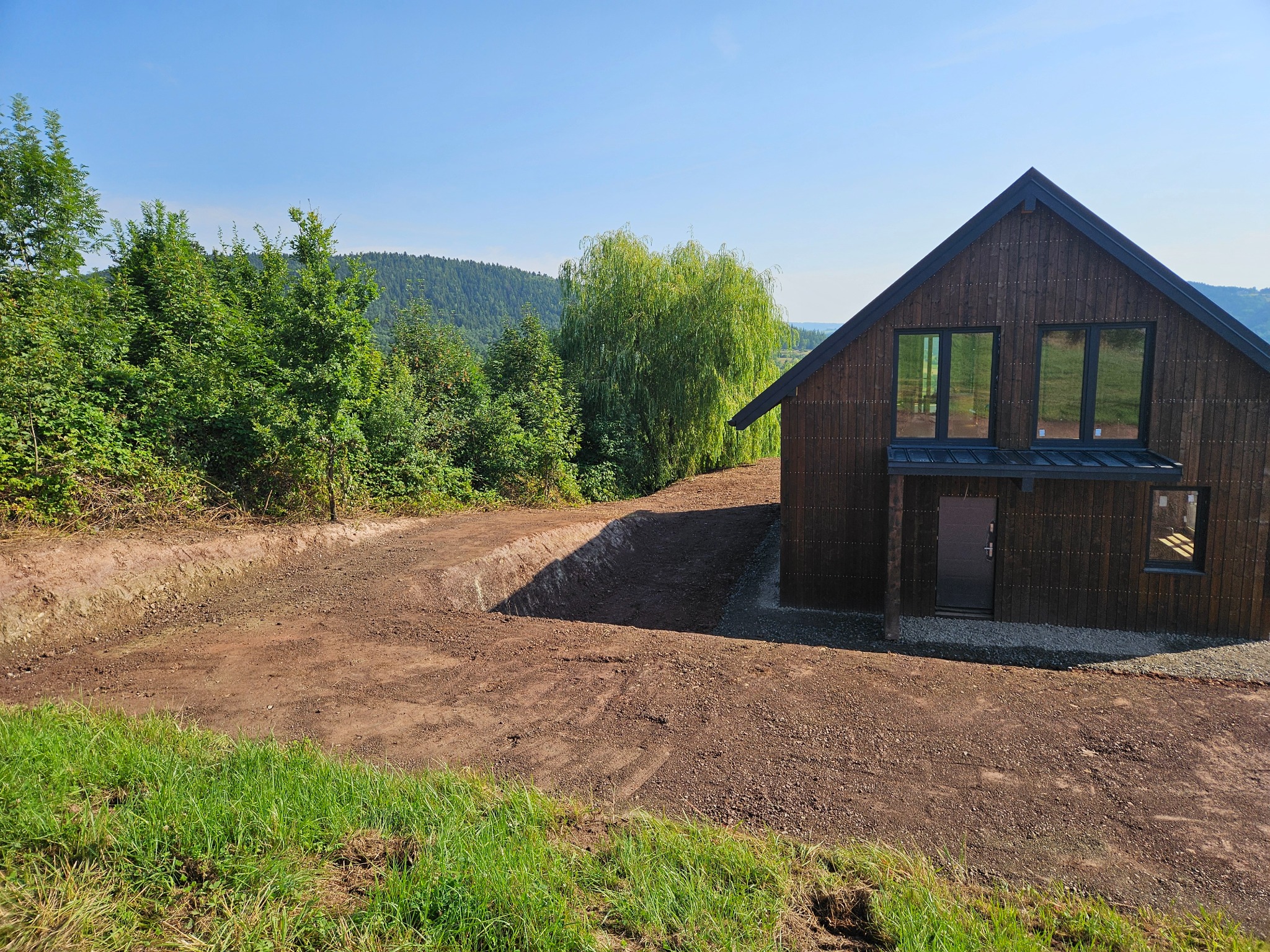 Przygotowany teren pod budowę domu z ciemnego drewna, widoczny wykop ziemny i wyrównany teren, w tle zieleń drzew i wzgórza, błękitne niebo.