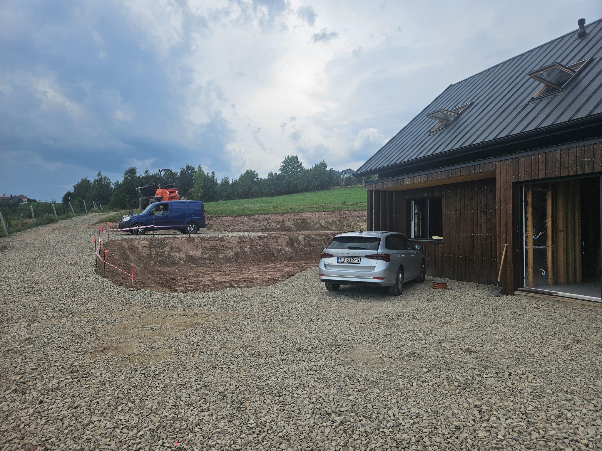 Teren budowy z częściowo utwardzoną drogą dojazdową, widoczny wykop, niebieski samochód dostawczy i traktor w tle, obok domu zaparkowany srebrny samochód osobowy.