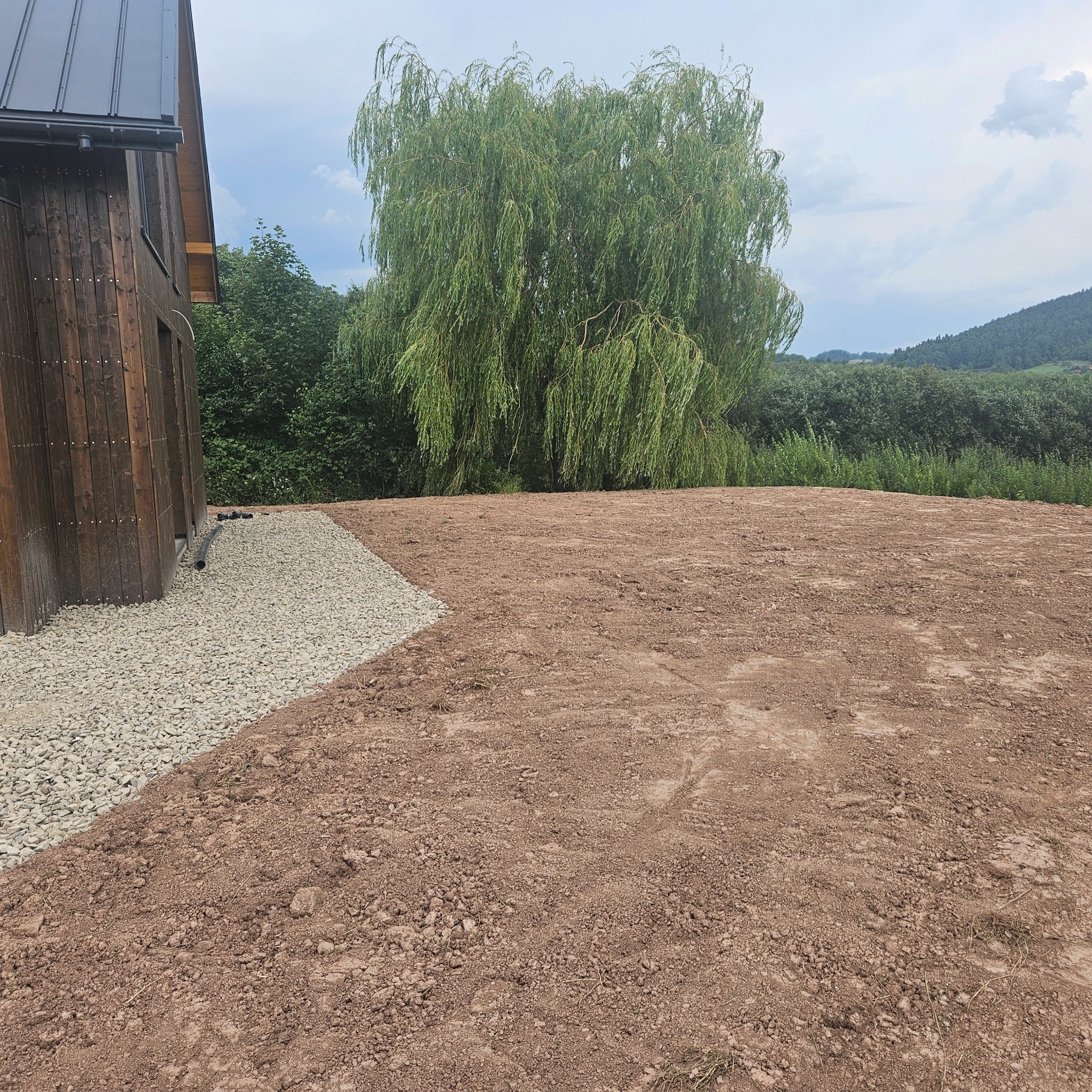 Przygotowany teren pod budowę domu z drewnianą elewacją, obok ułożony drenaż z kamieni, w tle drzewo i zieleń.