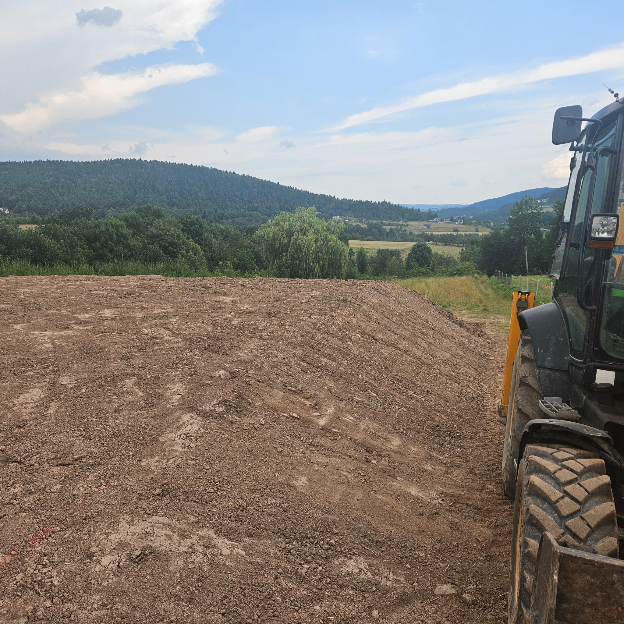 Przygotowany teren z widoczną stertą ziemi, częściowo widoczna koparka z boku, w tle zielone wzgórza i błękitne niebo.