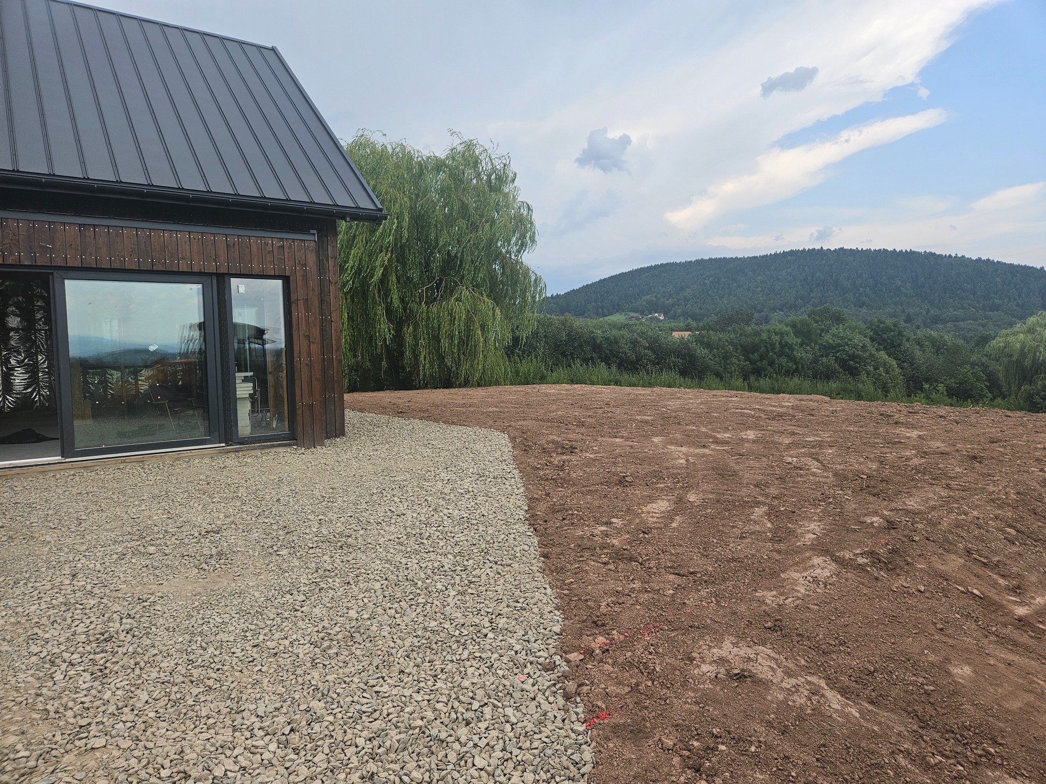 Przygotowany teren pod budowę domu jednorodzinnego: wyrównana ziemia granicząca z utwardzoną powierzchnią z kamienia, w tle widok na zalesione wzgórze i fragment nowoczesnego domu z ciemną elewacją.