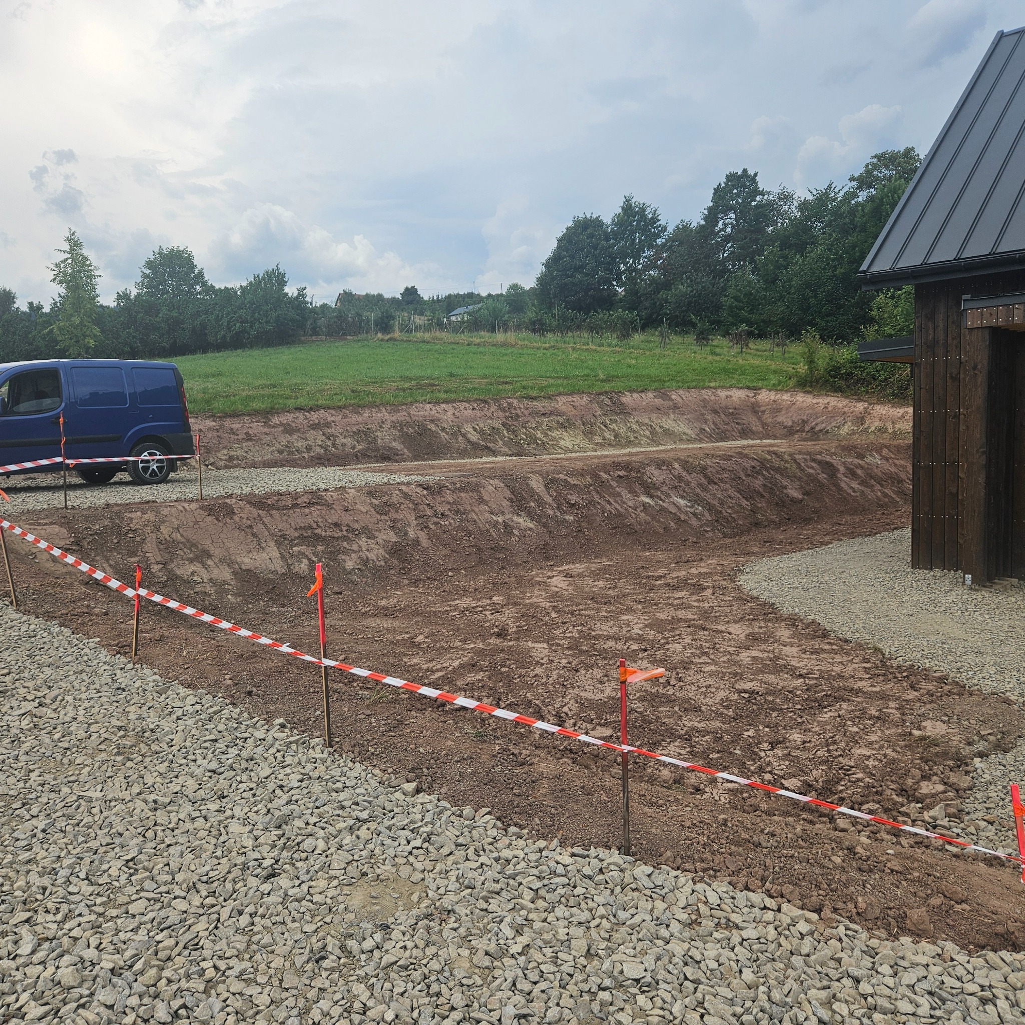 Przygotowany teren pod budowę z wykopem i częściowo utwardzoną drogą z boku budynku, zabezpieczony taśmą ostrzegawczą, z widocznym niebieskim busem w tle.