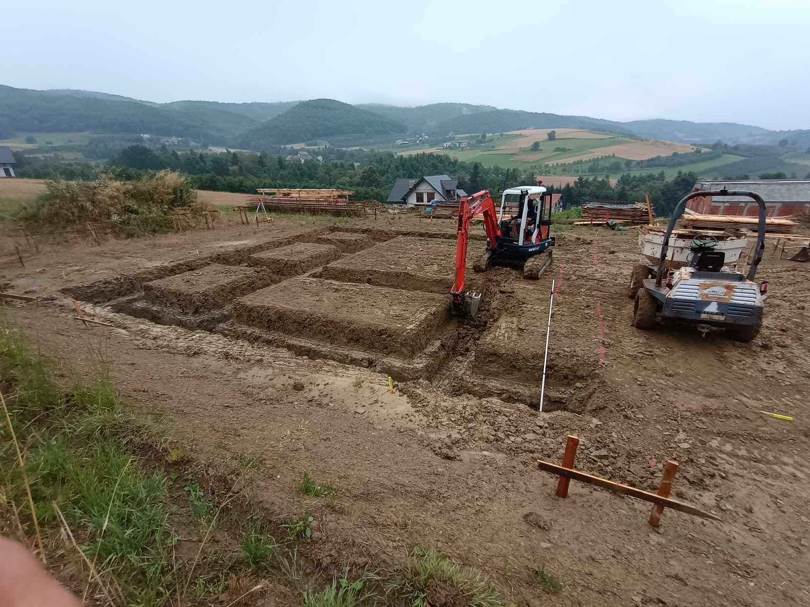 Wykop pod fundamenty domu jednorodzinnego z użyciem minikoparki Kubota i wywrotki, widoczne drewniane szalunki i tyczki geodezyjne, w tle pagórkowaty krajobraz.