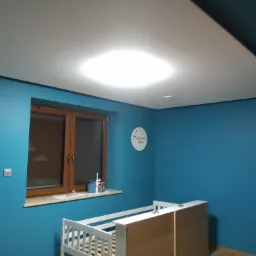 Pokój dziecięcy z turkusowymi ścianami, jasnym oknem i białym łóżeczkiem w trakcie montażu; widoczna okrągła dekoracja ścienna z napisem.