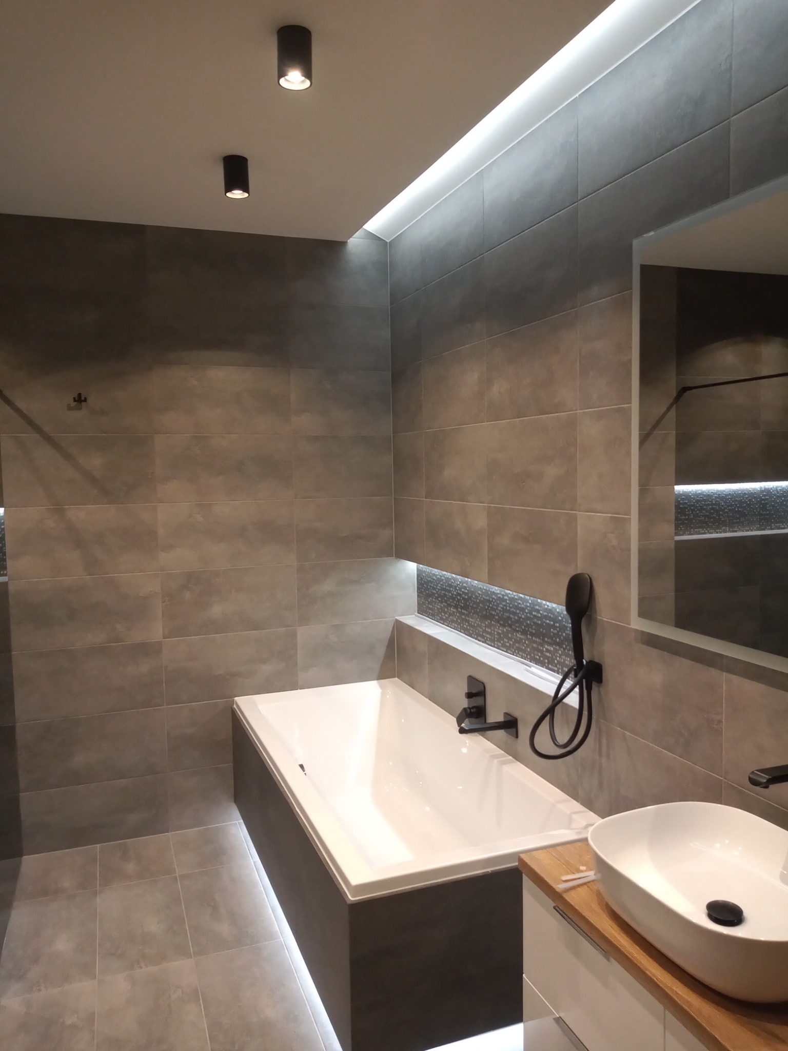 Nowoczesna łazienka w szarościach z podświetlaną wanną, umywalką nablatową na drewnianym blacie i czarną armaturą.