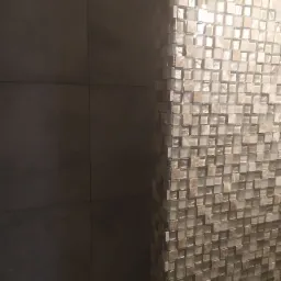 Fragment ściany wykończony mozaiką z małych, kwadratowych płytek w odcieniach beżu i bieli, sąsiadujący z gładką, szarą ścianą z dużych płytek.