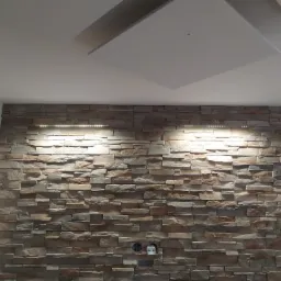 Ściana wykończona dekoracyjnym kamieniem z podświetleniem LED, widoczne gniazdka elektryczne, sufit z elementem dekoracyjnym.