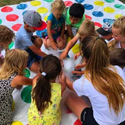 Grupa dzieci otacza białą parę na kolorowej macie do gry, niektóre dzieci dotykają dymu, a animator w białej koszulce nadzoruje zabawę.