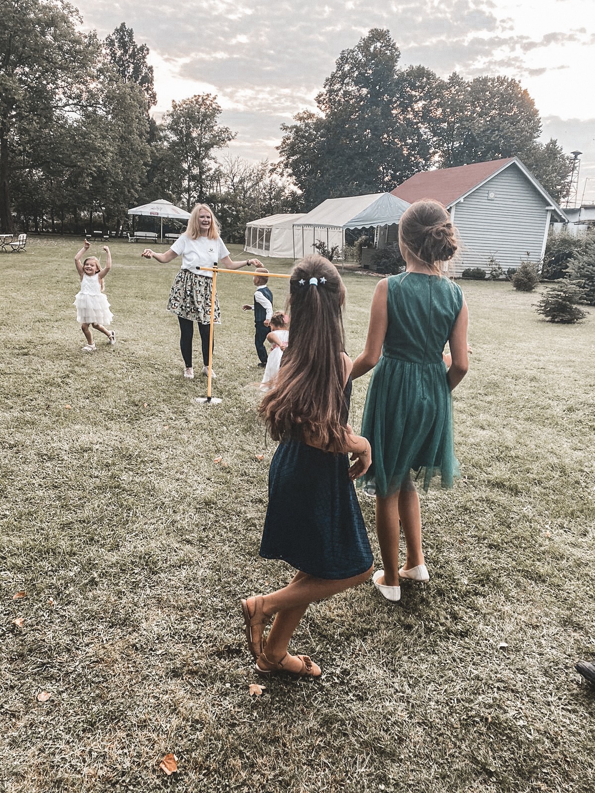 Na trawniku, kobieta z blond włosami trzyma żółtą poprzeczkę, pod którą przechodzą dzieci; w tle białe namioty i budynek.