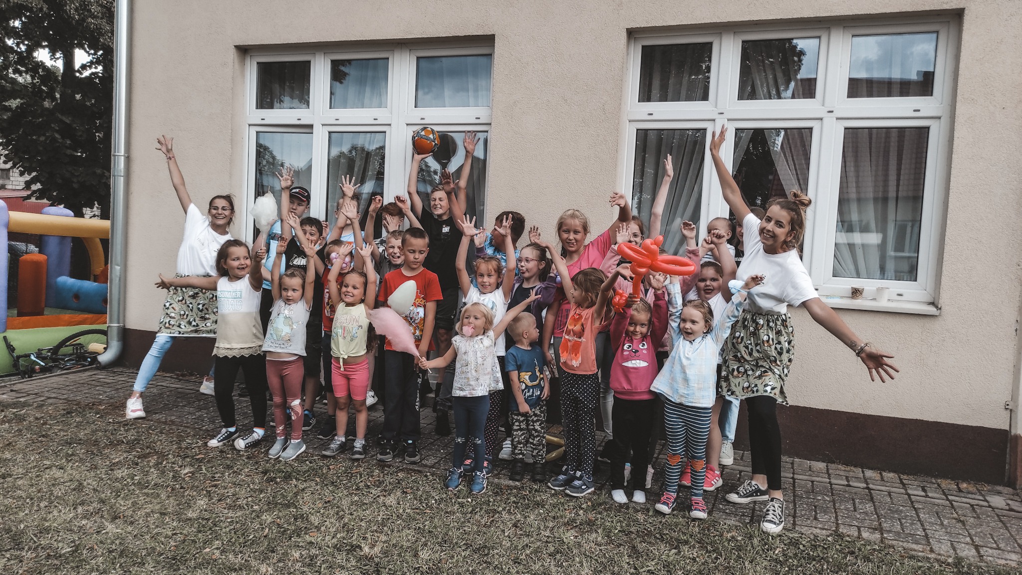 Grupa roześmianych dzieci i dwóch dorosłych pozuje z rękami w górze przed budynkiem z dwoma oknami, trzymając balon w kształcie serca, piłkę i watę cukrową.