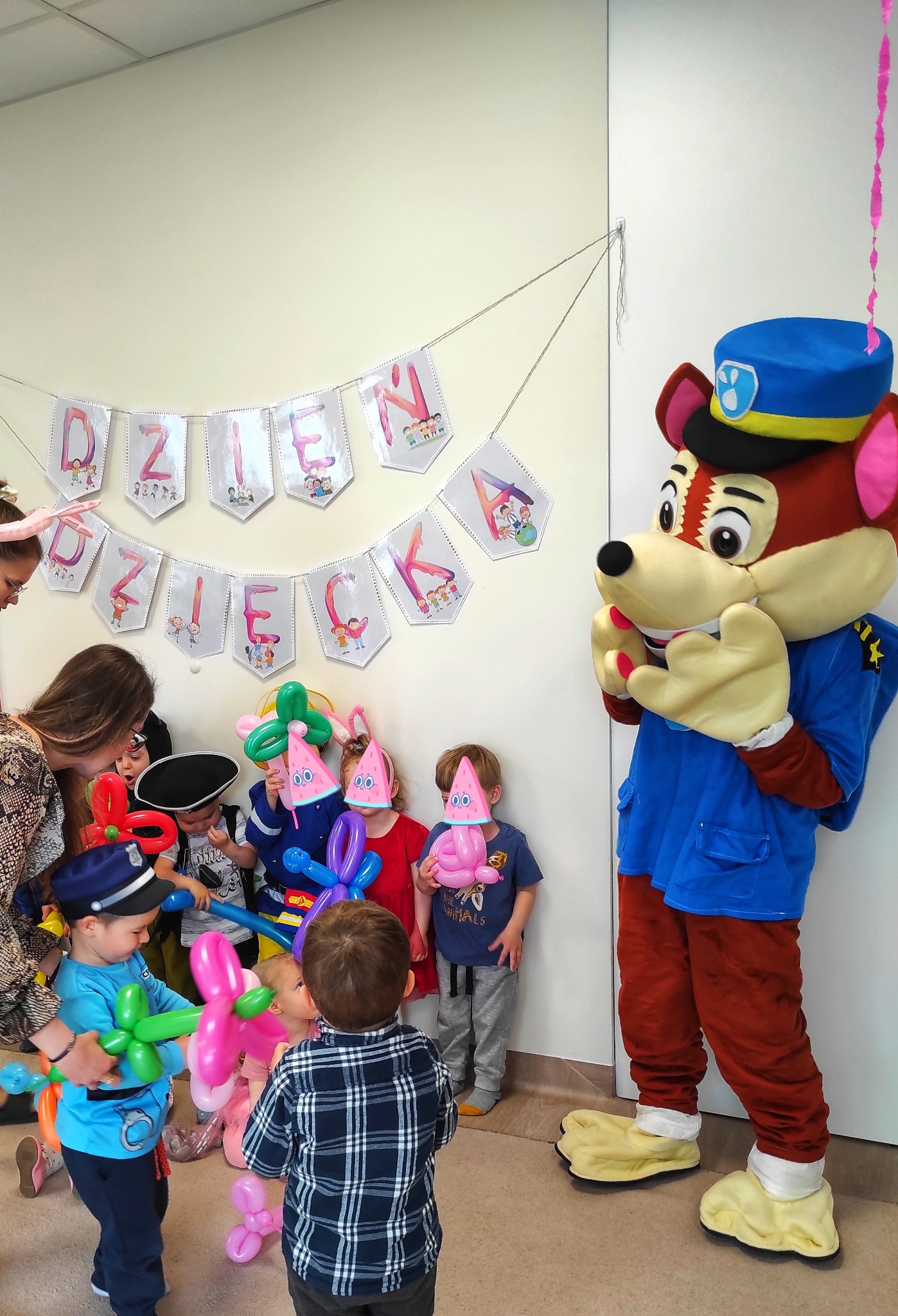 Dzieci z balonami modelowanymi w kształcie zwierząt i nakryć głowy, w towarzystwie osoby w kostiumie maskotki policyjnej, podczas imprezy urodzinowej z banerem 'Dzień Dziecka' w tle.