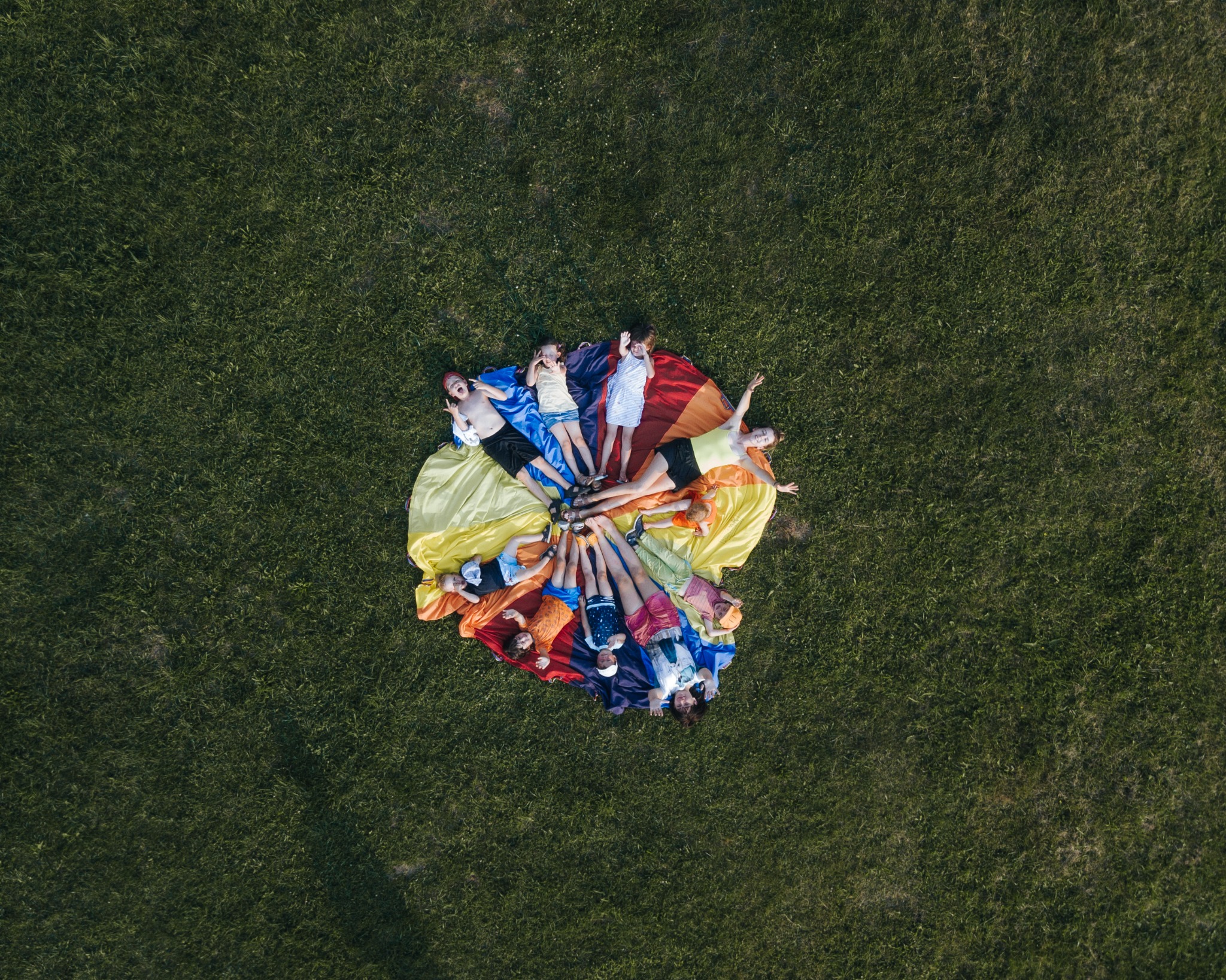 Widok z góry na grupę dzieci leżących na kolorowej chuście na trawie, tworzących okrąg.