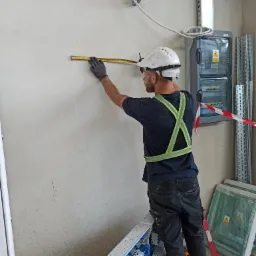 Elektryk w kasku i okularach ochronnych mierzy odległość na surowej ścianie za pomocą miarki, w tle skrzynka elektryczna i elementy instalacyjne