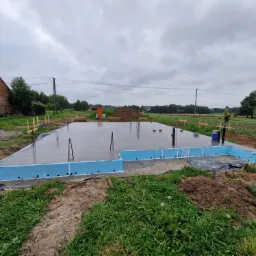 Wylany fundament z niebieską izolacją obwodową, wystającymi prętami zbrojeniowymi i rurami kanalizacyjnymi, widok na budynek gospodarczy i pole uprawne w tle, pochmurny dzień.