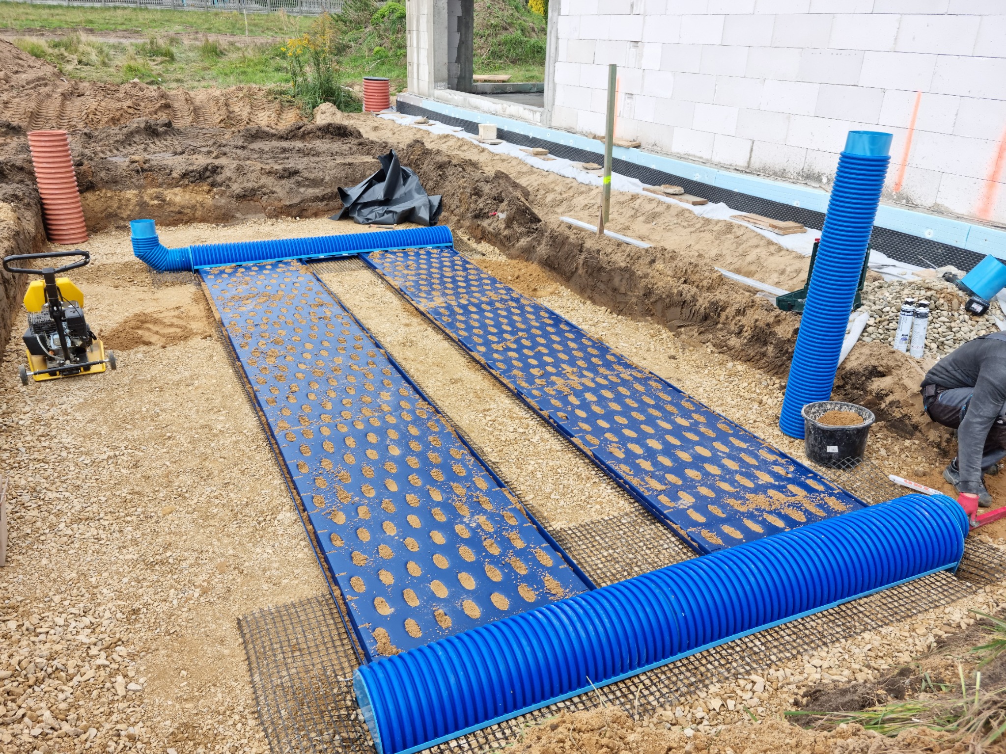 Przygotowanie podłoża pod budowę: ułożone niebieskie rury drenażowe z perforowanymi panelami na siatce, zagęszczarka w tle, widoczny fragment fundamentu budynku.
