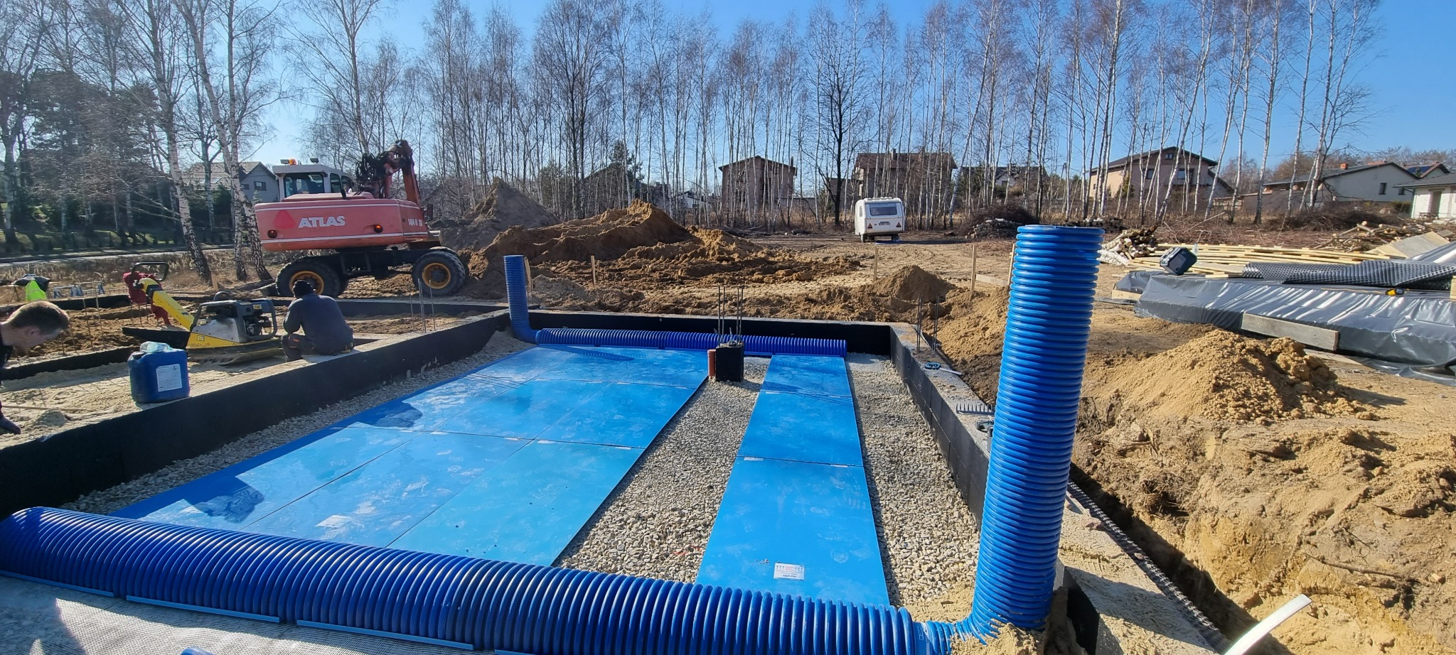 Przygotowanie podłoża pod płytę fundamentową z niebieską izolacją, drenażem z rur karbowanych i zagęszczarką w tle.