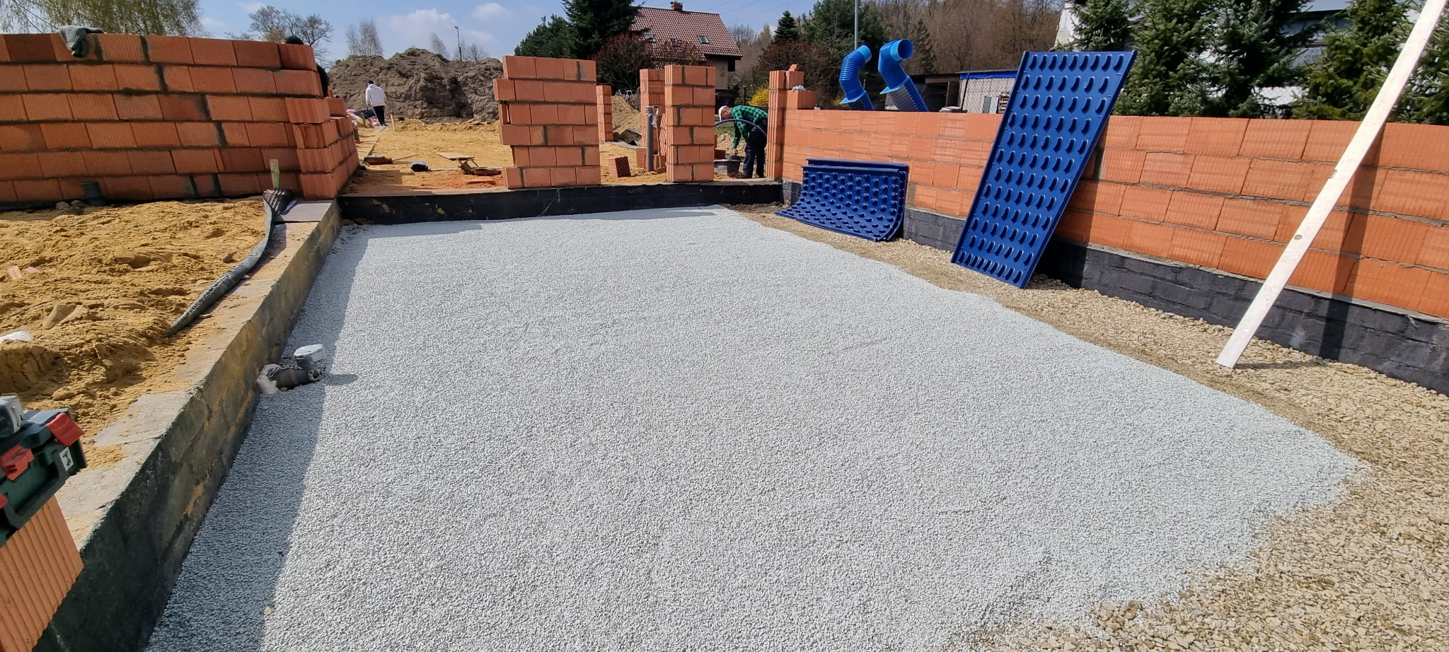 Wyrównany teren pod budowę z podsypką z grysu, widoczne ściany z cegieł dziurawki oraz niebieskie płyty izolacyjne.
