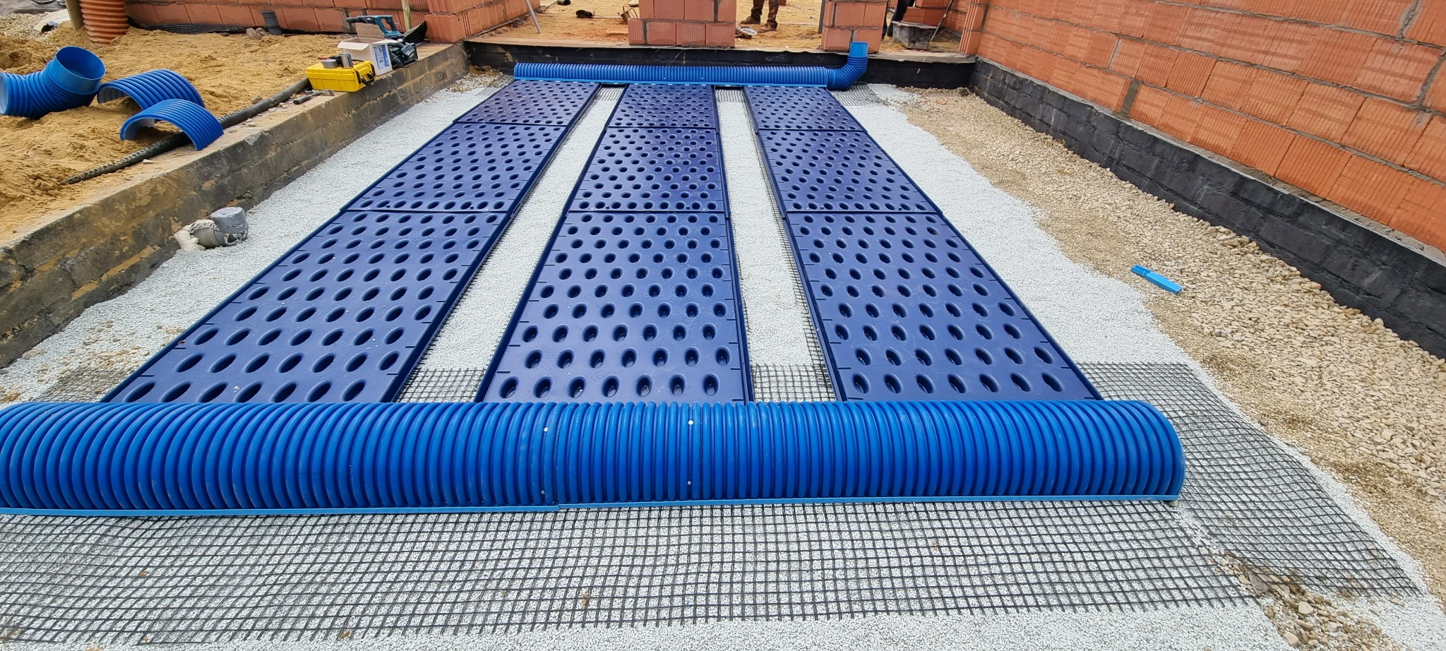 Instalacja drenażu: niebieskie rury karbowane i perforowane panele drenażowe ułożone na geowłókninie, obsypane żwirem przy fundamencie budynku z czerwonej cegły.