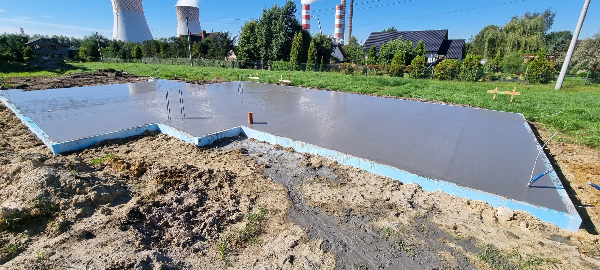 Wylany fundament z izolacją termiczną z niebieskiego styropianu, na tle elektrowni i zabudowań mieszkalnych, widoczne wystające pręty zbrojeniowe i pomarańczowa rura.