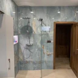 Wykończona łazienka z kabiną prysznicową typu walk-in, ściany wyłożone turkusowymi płytkami imitującymi marmur, beżowa podłoga z płytek oraz drewniane drzwi.