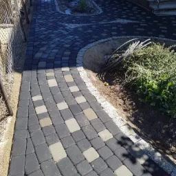 Ułożona kostka brukowa w odcieniach szarości i grafitu, z jasnymi akcentami, tworząca ścieżkę z obrzeżem z kostki granitowej, widok z góry, częściowo zacieniona.