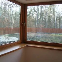 Drewniane okno narożne z widokiem na ogród i las w tle; widoczny fragment parapetu z zaokrąglonym brzegiem