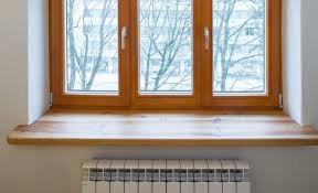 Jasne drewniane okno z trzema skrzydłami i szerokim parapetem, poniżej biały grzejnik panelowy, widok na drzewa bez liści za oknem, ściana w neutralnym odcieniu.