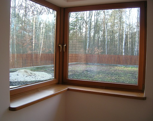 Drewniane okno narożne z widokiem na ogród i las w tle; widoczny fragment parapetu z zaokrąglonym brzegiem