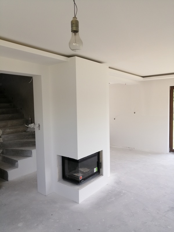 Surowe wnętrze w trakcie wykończenia: białe ściany po tynkowaniu, zabudowa kominkowa z wkładem, schody w stanie surowym, wisząca żarówka bez klosza.