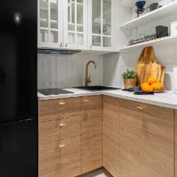 Kąt ujęcia na jasną kuchnię z drewnianymi szafkami, złotą armaturą i czarną lod&oacute;wką. Na blacie owoce i deska. Białe płytki na ścianie i otwarte p&oacute;łki.