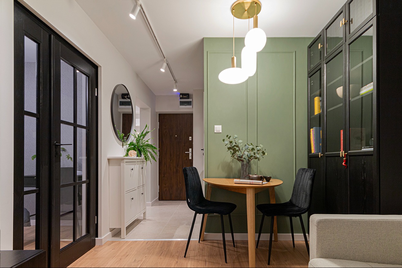 Nowoczesne wykończenie wnętrza: czarne drzwi z przeszkleniami, okrągłe lustro i zielona ściana z okrągłym stolikiem i czarnymi krzesłami. Oświetlenie punktowe sufitowe.