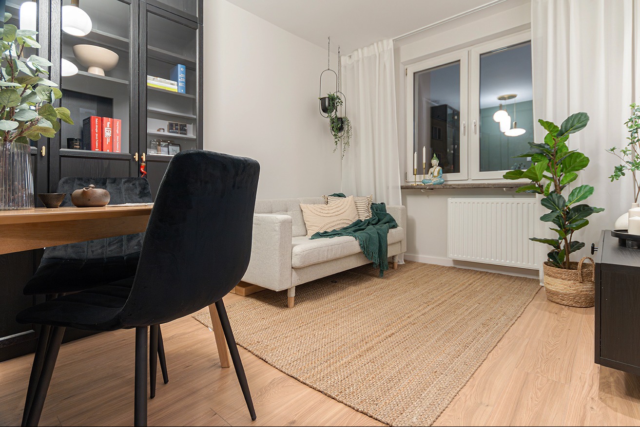 Jasne wnętrze: sofa z poduszkami i pledem, dywan z juty, okno z zasłonami, czarne krzesła przy drewnianym stole. Rośliny doniczkowe dodają przytulności.