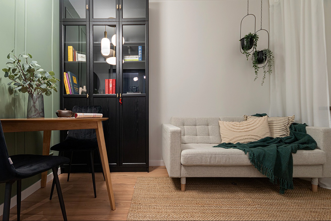 Jasny salon z drewnianym stołem, czarnym regałem z przeszklonymi drzwiami i szarą sofą z zielonym pledem, dopełniony wiszącymi roślinami i dywanem z juty.