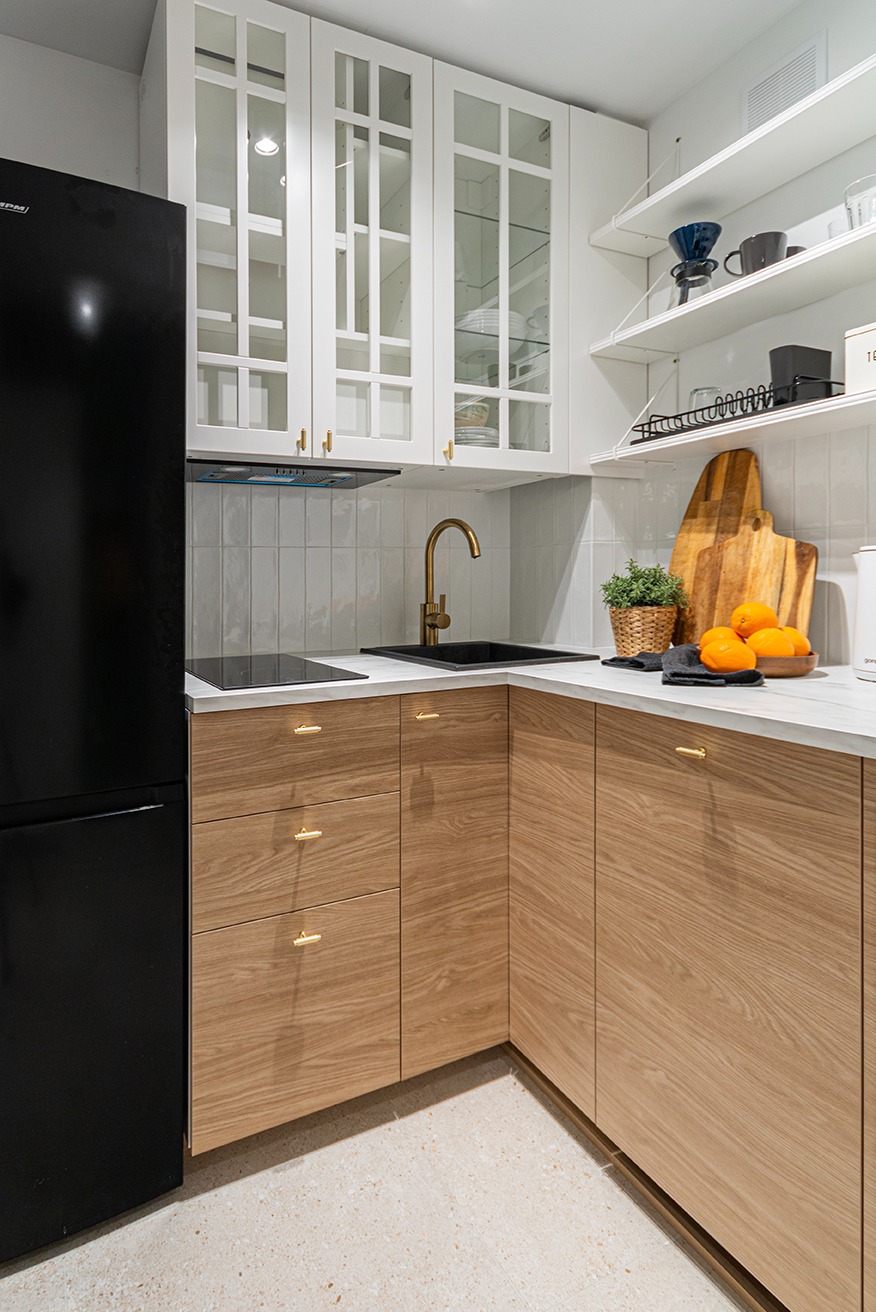 Kąt ujęcia na jasną kuchnię z drewnianymi szafkami, złotą armaturą i czarną lodówką. Na blacie owoce i deska. Białe płytki na ścianie i otwarte półki.