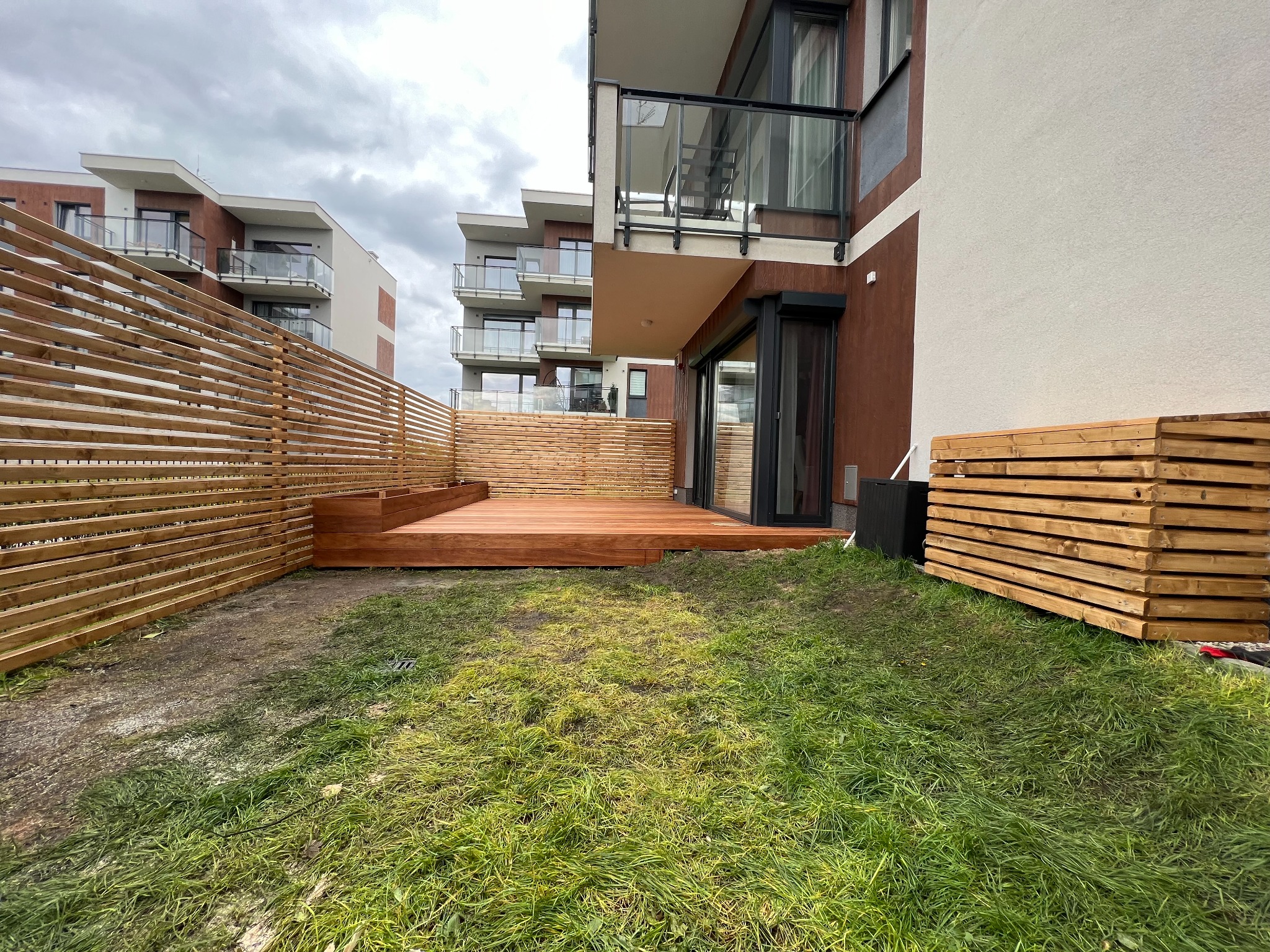 Nowoczesny taras drewniany z ażurowym ogrodzeniem, przylegający do budynku z balkonami. Widoczna trawa i elementy drewniane. Pochmurne niebo w tle.