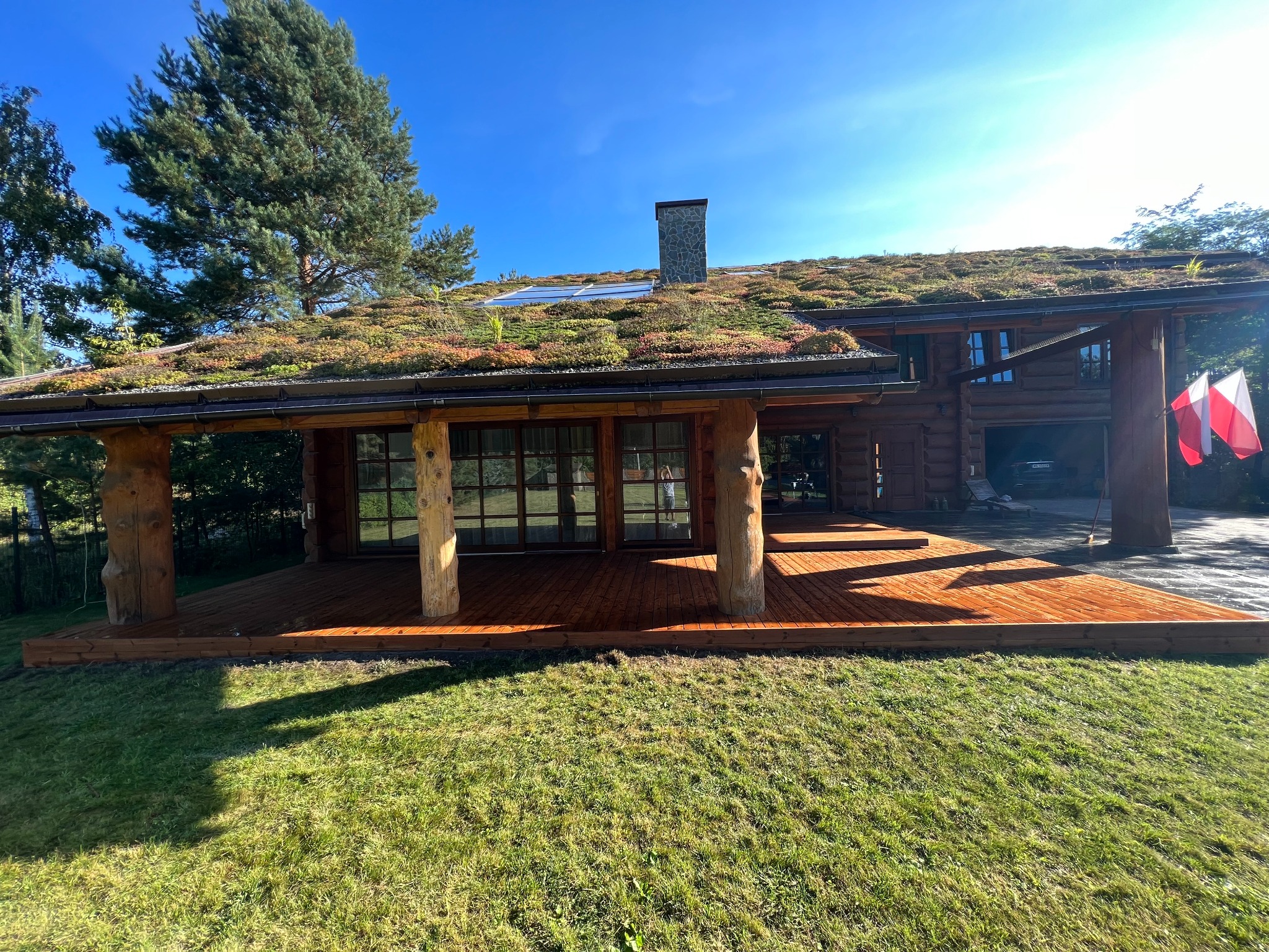 Drewniany dom z zielonym dachem i tarasem. Widoczne drewniane kolumny, polska flaga i trawnik. Dach pokryty roślinnością, z kominem i panelami słonecznymi.